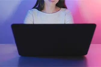 A woman seated at a table, working on a laptop with a camera positioned nearby.