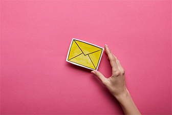  A hand grips a bright yellow envelope, set against a soft pink backdrop, highlighting the envelope's color.