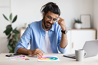 A man with glasses and a beard sits at a table, working on a laptop beside a color wheel.