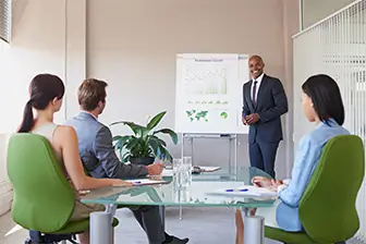 A man in a suit delivers a presentation to an engaged audience of colleagues in a professional setting.