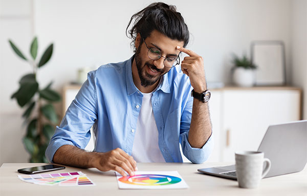 A man with glasses and a beard sits at a table, working on a laptop beside a color wheel.