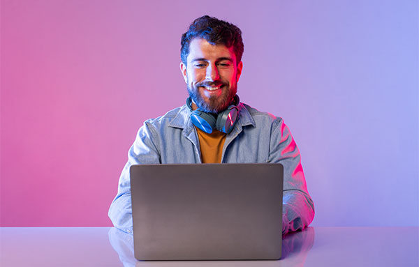 A smiling man with a beard wears headphones while using a laptop, showcasing a moment of enjoyment and focus.